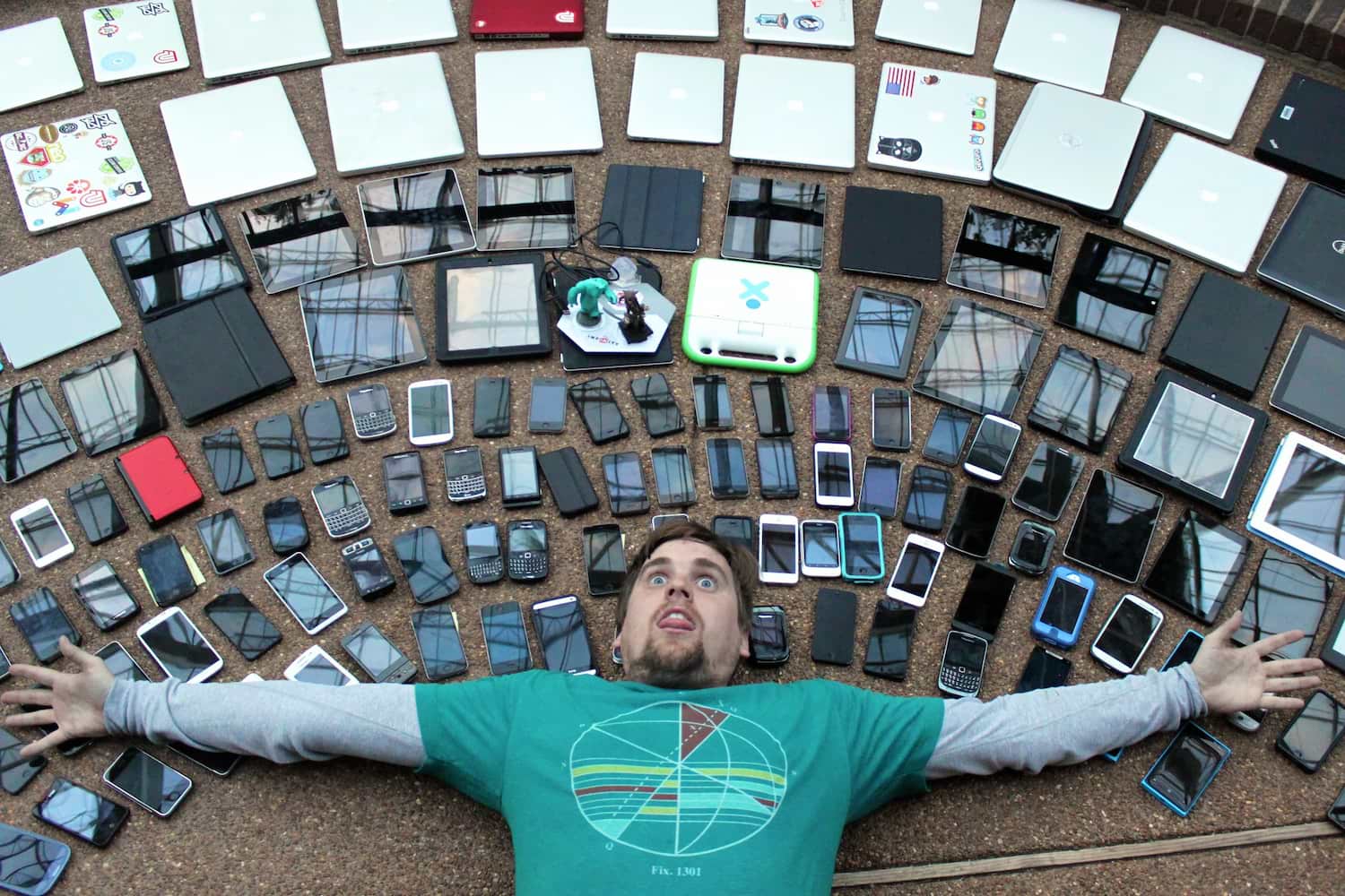 A man lies on the floor, arms stretched, as if he's been knocked out. Above him, arranged in concentric semi-circles are many different kinds of phones and tablets.