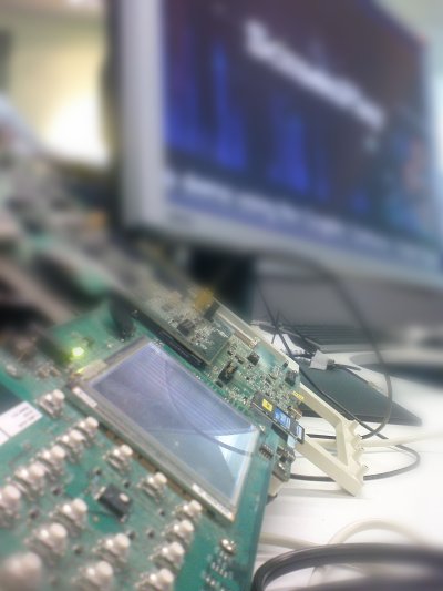 Close-up photo of a development board with a monitor visible in the background. The monitor is out of focus, so the contents of the screen cannot be seen clearly.