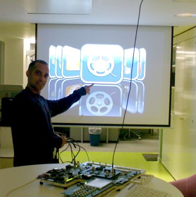 Joseph stands behind a development board and points at a projector screen in the background on which the demo's UI is visible