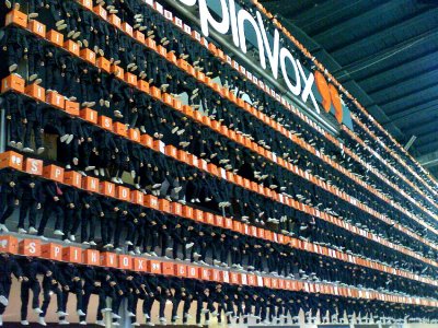 A wall made up of hundreds of toy figurines in black suits with bright orange cubes for heads. Each cube has a white letter, and together they spell out words like "Spinvox".